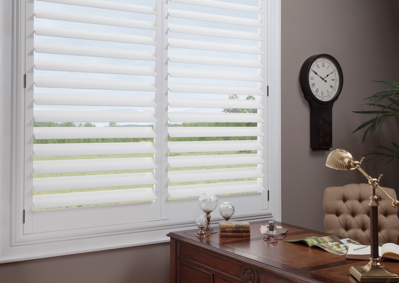 NewStyle® Hybrid Shutters in a home office with a warm wood desk, brass lamp, and white hybrid shutter panels