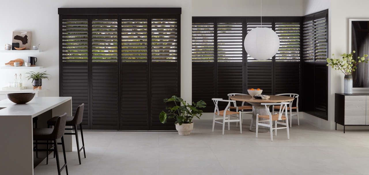Heritance® Hardwood Shutters across a large dining and kitchen opening with dark richly finished panels
